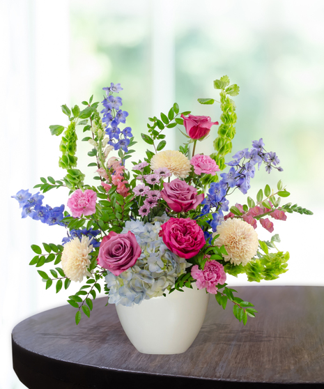 Malibu Splash flower arrangement featuring magenta garden roses, lavender roses, blue hydrangeas, and blue delphinium in a matte white ceramic bowl.