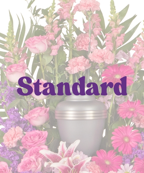 Feminine Cremation Arrangement featuring pink roses, carnations, gerbera daisies, rose lilies, purple stock, and tropical foliage surrounding a gray urn
