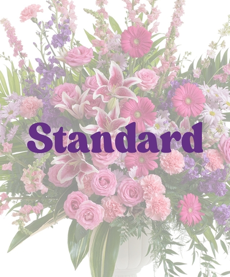 Feminine urn floral arrangement with pink rose lilies, roses, carnations, gerbera daisies, purple stock, and greenery in a white pedestal vase