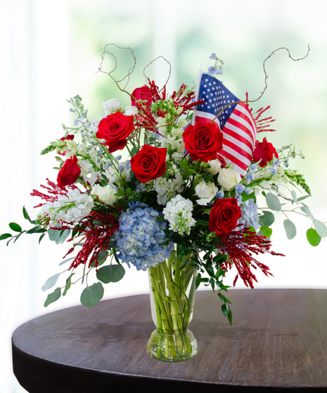 Rocket's Red Glare Independence Day flower arrangement featuring red roses, blue hydrangeas, white stock, and sparkly red firework accents in a clear glass vase.