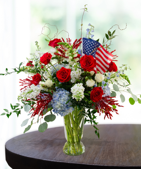 Rocket's Red Glare Independence Day flower arrangement featuring red roses, blue hydrangeas, white stock, and sparkly red firework accents in a clear glass vase.