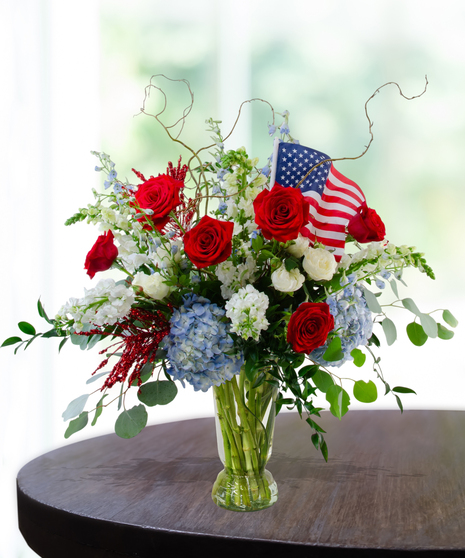Rocket's Red Glare Independence Day flower arrangement featuring red roses, blue hydrangeas, white stock, and sparkly red firework accents in a clear glass vase.