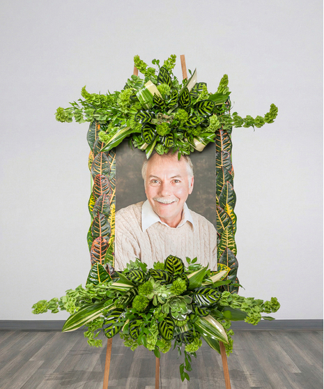 Floral photo frame tribute with croton leaves, green foliage, and tropical accents on an easel from Billy Heroman’s in Baton Rouge