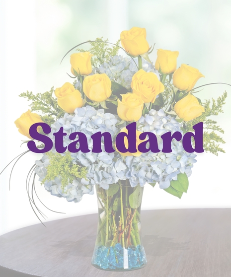Southern Pride collegiate floral arrangement featuring yellow roses, light blue hydrangeas, golden solidago, and bear grass in a clear glass vase with blue decorative gems.