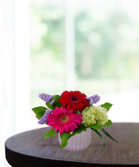 Grateful for You floral arrangement featuring magenta and blush gerbera daisies, pink carnations, and purple statice in a textured lavender glass vase.