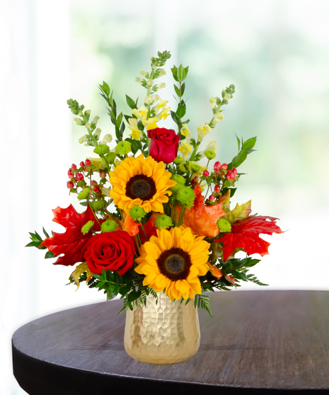 Fall flower arrangement with sunflowers, red roses, snapdragons, berries, and autumn leaves in a gold hammered vase.