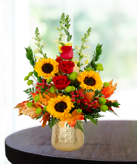 Fall flower arrangement with sunflowers, red roses, snapdragons, berries, and autumn leaves in a gold hammered vase.
