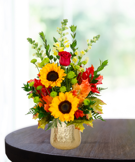 Fall flower arrangement with sunflowers, red roses, snapdragons, berries, and autumn leaves in a gold hammered vase.