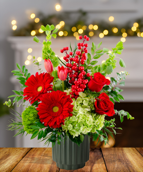 Deck the Halls vibrant Christmas arrangement with red gerbera daisies, red roses, pink tulips, green hydrangeas, Bells of Ireland, and red berries in a green textured vase