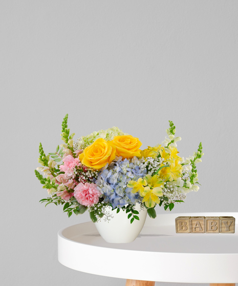 Heaven Sent flower arrangement with yellow roses, blue hydrangeas, pink carnations, and alstroemeria in a cream pedestal vase for a baby shower or gender-neutral gift
