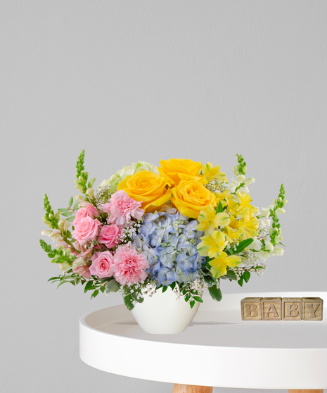 Heaven Sent flower arrangement with yellow roses, blue hydrangeas, pink carnations, and alstroemeria in a cream pedestal vase for a baby shower or gender-neutral gift