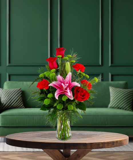 Secret Santa Christmas flower arrangement with red roses, pink rose lilies, red mini carnations, green button mums, and evergreen in a clear glass vase