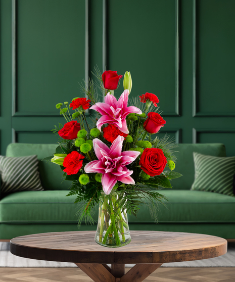 Secret Santa Christmas flower arrangement with red roses, pink rose lilies, red mini carnations, green button mums, and evergreen in a clear glass vase