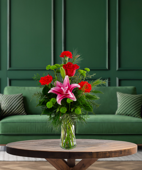 Secret Santa Christmas flower arrangement with red roses, pink rose lilies, red mini carnations, green button mums, and evergreen in a clear glass vase