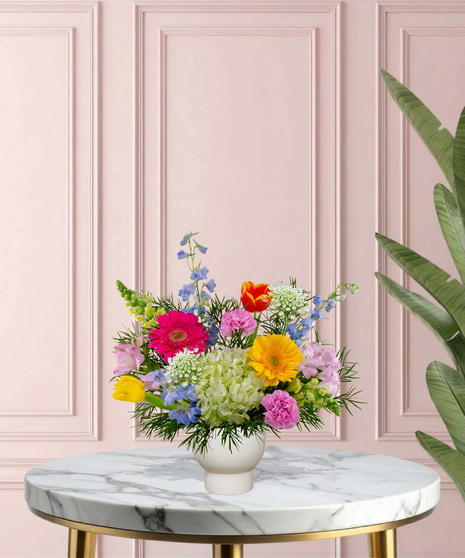 She Knows Best floral arrangement featuring coral garden roses, vibrant gerbera daisies, blue delphinium, and lime hydrangeas in a white pedestal vase.