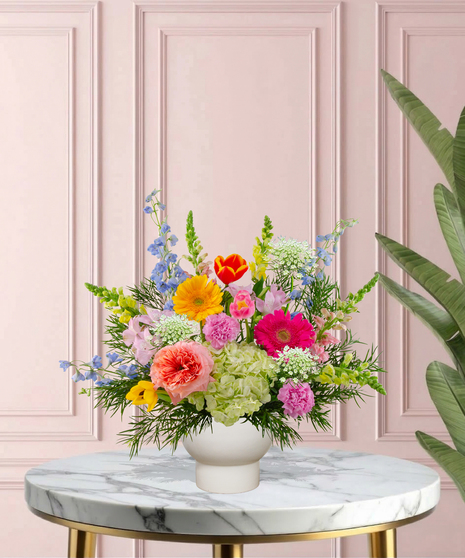 She Knows Best floral arrangement featuring coral garden roses, vibrant gerbera daisies, blue delphinium, and lime hydrangeas in a white pedestal vase.