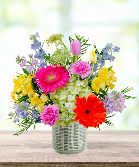 Hummingbird Hues floral arrangement with peach garden roses, green hydrangeas, gerbera daisies, and mixed colorful blooms in a blue-and-white patterned vase on a wooden table.
