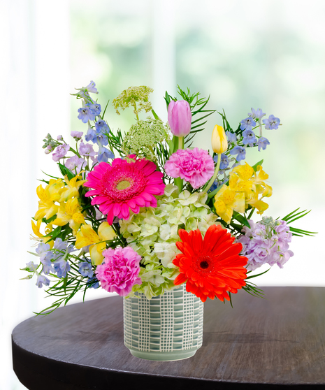 Hummingbird Hues floral arrangement with peach garden roses, green hydrangeas, gerbera daisies, and mixed colorful blooms in a blue-and-white patterned vase on a wooden table.