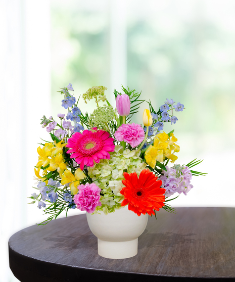 Hummingbird Hues floral arrangement with peach garden roses, green hydrangeas, gerbera daisies, and mixed colorful blooms in a white pedestal vase on a wooden table.