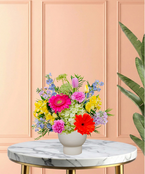 Hummingbird Hues floral arrangement with peach garden roses, green hydrangeas, gerbera daisies, and mixed colorful blooms in a blue-and-white patterned vase on a wooden table.