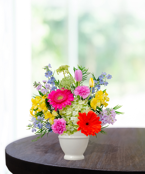 Hummingbird Hues floral arrangement with peach garden roses, green hydrangeas, gerbera daisies, and mixed colorful blooms in a white pedestal vase on a wooden table.