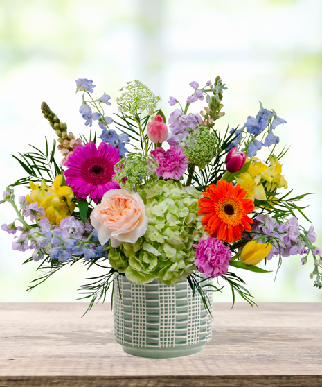 Hummingbird Hues floral arrangement with peach garden roses, green hydrangeas, gerbera daisies, and mixed colorful blooms in a blue-and-white patterned vase on a wooden table.