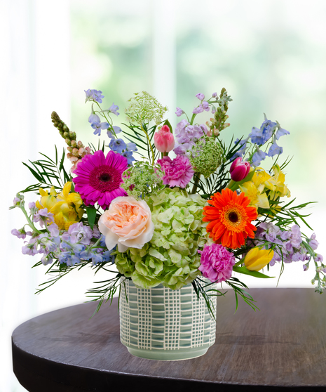 Hummingbird Hues floral arrangement with peach garden roses, green hydrangeas, gerbera daisies, and mixed colorful blooms in a blue-and-white patterned vase on a wooden table.