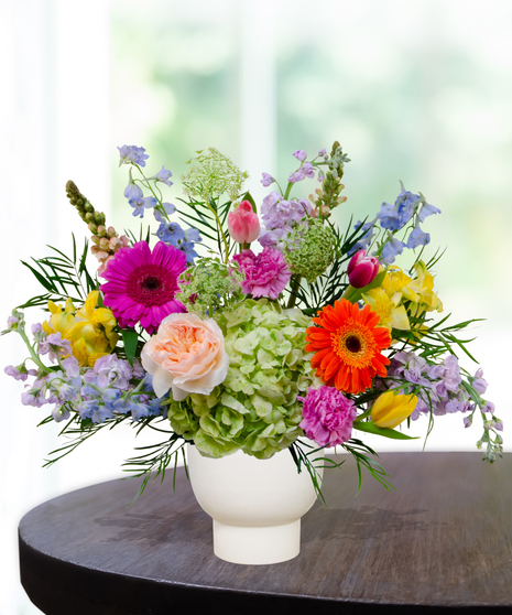 Hummingbird Hues floral arrangement with peach garden roses, green hydrangeas, gerbera daisies, and mixed colorful blooms in a white pedestal vase on a wooden table.