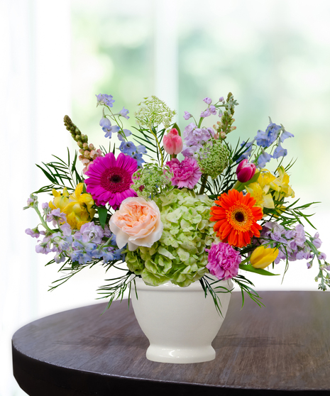 Hummingbird Hues floral arrangement with peach garden roses, green hydrangeas, gerbera daisies, and mixed colorful blooms in a white pedestal vase on a wooden table.
