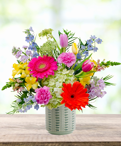 Hummingbird Hues floral arrangement with peach garden roses, green hydrangeas, gerbera daisies, and mixed colorful blooms in a blue-and-white patterned vase on a wooden table.