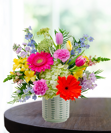 Hummingbird Hues floral arrangement with peach garden roses, green hydrangeas, gerbera daisies, and mixed colorful blooms in a blue-and-white patterned vase on a wooden table.