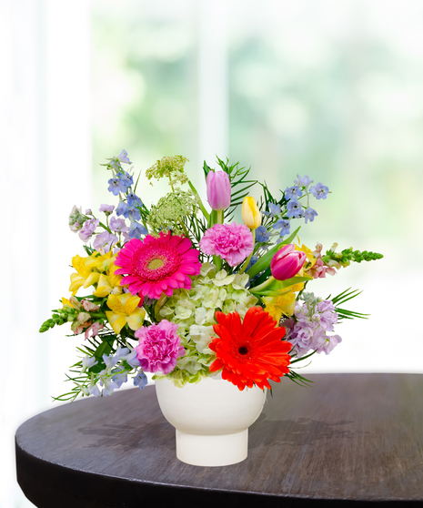 Hummingbird Hues floral arrangement with peach garden roses, green hydrangeas, gerbera daisies, and mixed colorful blooms in a white pedestal vase on a wooden table.