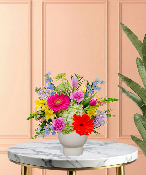 Hummingbird Hues floral arrangement with peach garden roses, green hydrangeas, gerbera daisies, and mixed colorful blooms in a blue-and-white patterned vase on a wooden table.