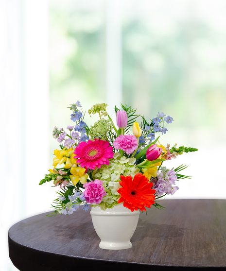 Hummingbird Hues floral arrangement with peach garden roses, green hydrangeas, gerbera daisies, and mixed colorful blooms in a white pedestal vase on a wooden table.