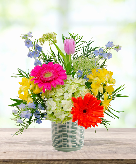 Hummingbird Hues floral arrangement with peach garden roses, green hydrangeas, gerbera daisies, and mixed colorful blooms in a blue-and-white patterned vase on a wooden table.