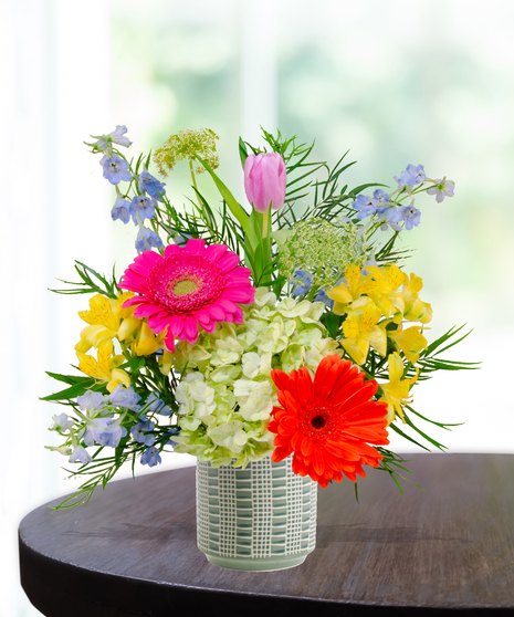 Hummingbird Hues floral arrangement with peach garden roses, green hydrangeas, gerbera daisies, and mixed colorful blooms in a blue-and-white patterned vase on a wooden table.