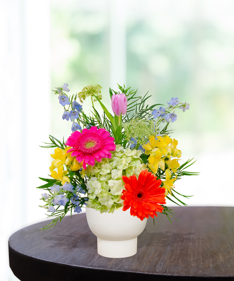 Hummingbird Hues floral arrangement with peach garden roses, green hydrangeas, gerbera daisies, and mixed colorful blooms in a white pedestal vase on a wooden table.