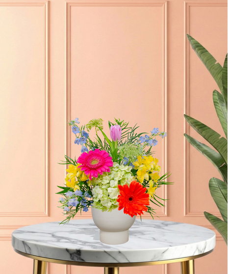 Hummingbird Hues floral arrangement with peach garden roses, green hydrangeas, gerbera daisies, and mixed colorful blooms in a blue-and-white patterned vase on a wooden table.