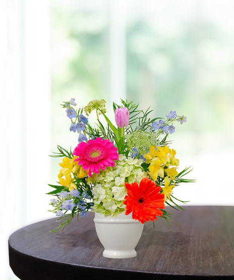 Hummingbird Hues floral arrangement with peach garden roses, green hydrangeas, gerbera daisies, and mixed colorful blooms in a white pedestal vase on a wooden table.