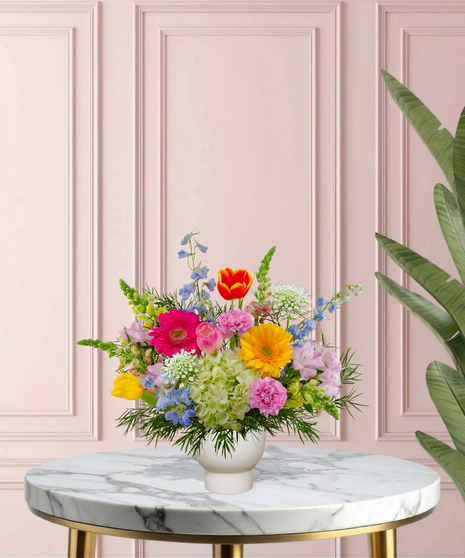 She Knows Best floral arrangement featuring coral garden roses, vibrant gerbera daisies, blue delphinium, and lime hydrangeas in a white pedestal vase.