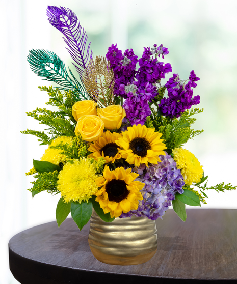 A festive Mardi Gras flower arrangement with yellow roses, sunflowers, purple stock, hydrangeas, and glittering peacock feathers in a gold vase.