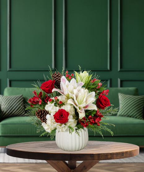 Jolly Jingle Berry Christmas arrangement with white lilies, red roses, white hydrangeas, red alstroemeria, red berries, pine cones, and evergreen in a textured white vase