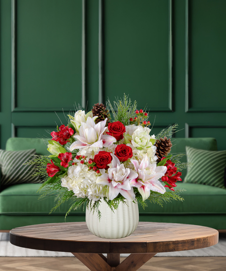 Jolly Jingle Berry Christmas arrangement with white lilies, red roses, white hydrangeas, red alstroemeria, red berries, pine cones, and evergreen in a textured white vase