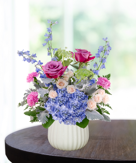 Sweet June flower arrangement in a white vase featuring pink roses, blue hydrangeas, lavender liatris, and delphinium