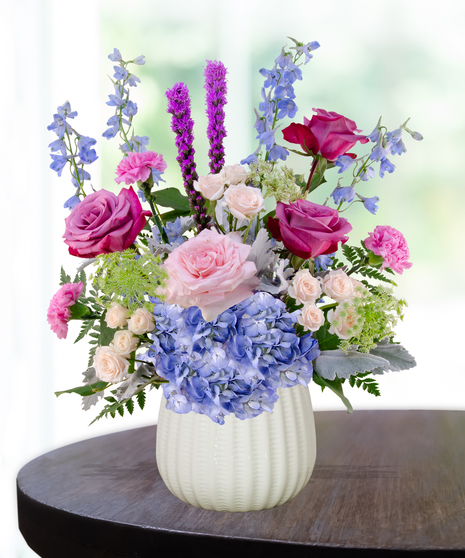 Sweet June flower arrangement in a white vase featuring pink roses, blue hydrangeas, lavender liatris, and delphinium