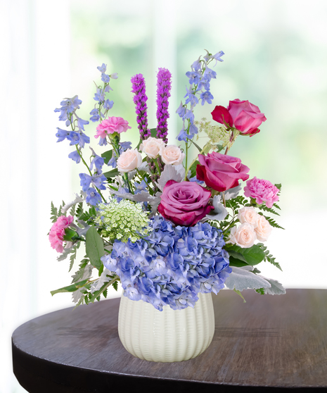 Sweet June flower arrangement in a white vase featuring pink roses, blue hydrangeas, lavender liatris, and delphinium