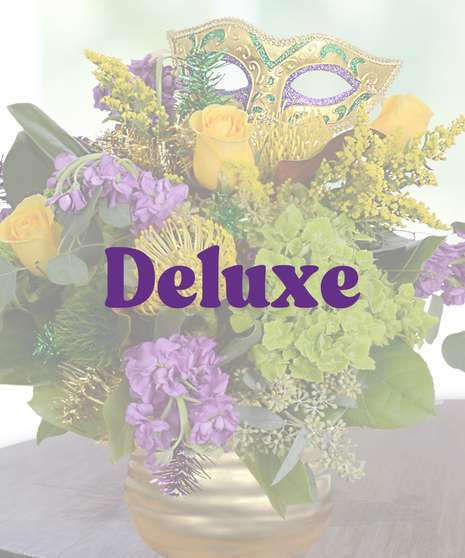 A luxurious Mardi Gras-themed flower arrangement featuring yellow roses, purple stock flowers, green hydrangeas, pincushion protea, and a festive mask in a gold vase.