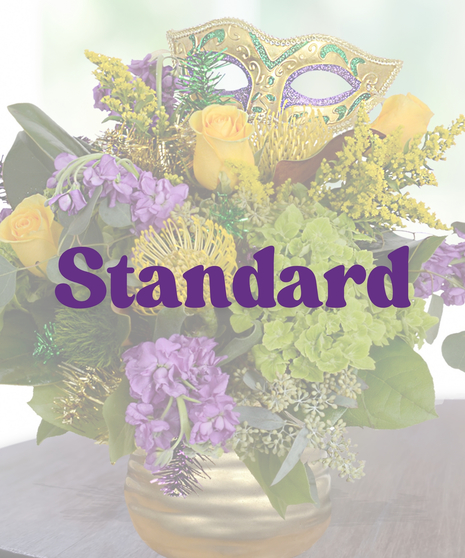 A luxurious Mardi Gras-themed flower arrangement featuring yellow roses, purple stock flowers, green hydrangeas, pincushion protea, and a festive mask in a gold vase.