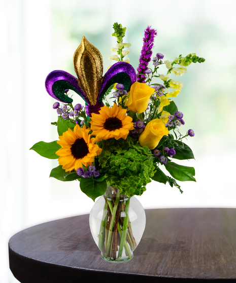Mardi Gras floral arrangement with sunflowers, yellow roses, and a glittery Fleur de Lis insert in a glass vase.