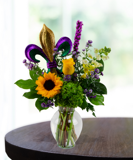 Mardi Gras floral arrangement with sunflowers, yellow roses, and a glittery Fleur de Lis insert in a glass vase.